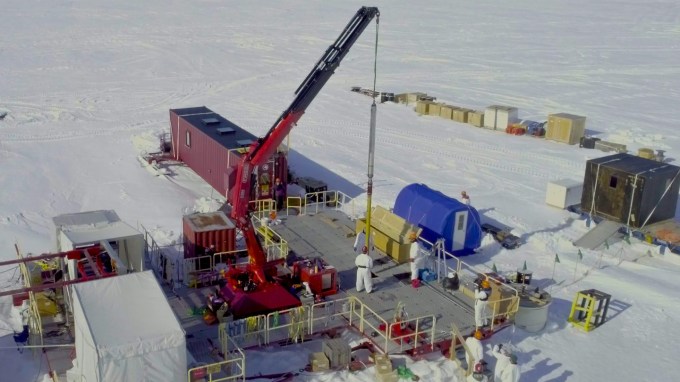 A research team in white protective suits works on a platform in Antarctica with a red crane lowering equipment into the ice. Portable shelters and supply containers surround the site.