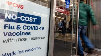 A person wearing a green jacket and blue pants walks into a CVS. Shown in the store window to the person's left is a sign advertising "No-cost flu and COVID-19 vaccines with most insurance. Walk-ins welcome"