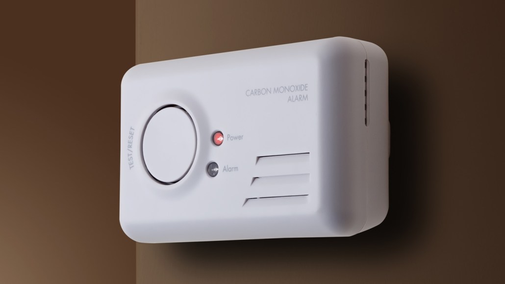 A white carbon monoxide monitor is shown mounted on a dark brown wall. The alarm has a large white button to "test/reset" on the left. To the right of the button are two round indicators for "power" and "alarm"