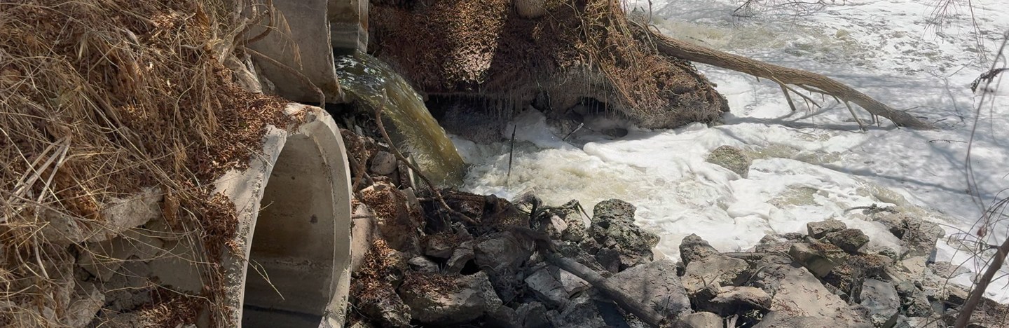 River water is shown rapidly flowing through concrete culverts, creating foam in the Tijuana River. The water appears brownish black because it is contaminated with untreated sewage.