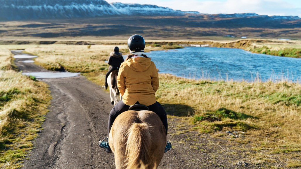 Two people riding horses by an alpine pond are shown from behind.