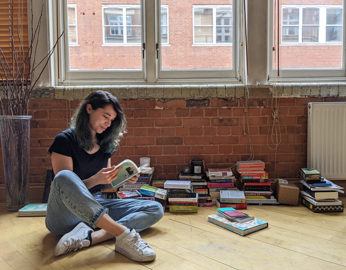 A woman sits cross-legged on the floor looking through a book. Piles of other books are behind her.