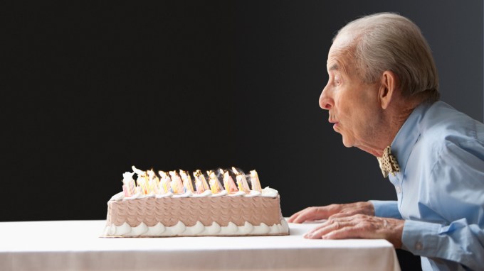 An elderly man in a blue collared shirt and bowtie leans over the edge of a table to blow out dozens of candles on a birthday cake. Elderly, healthy adults might benefit from brain mapping to detect early signs of depression and Alzheimer’s disease.