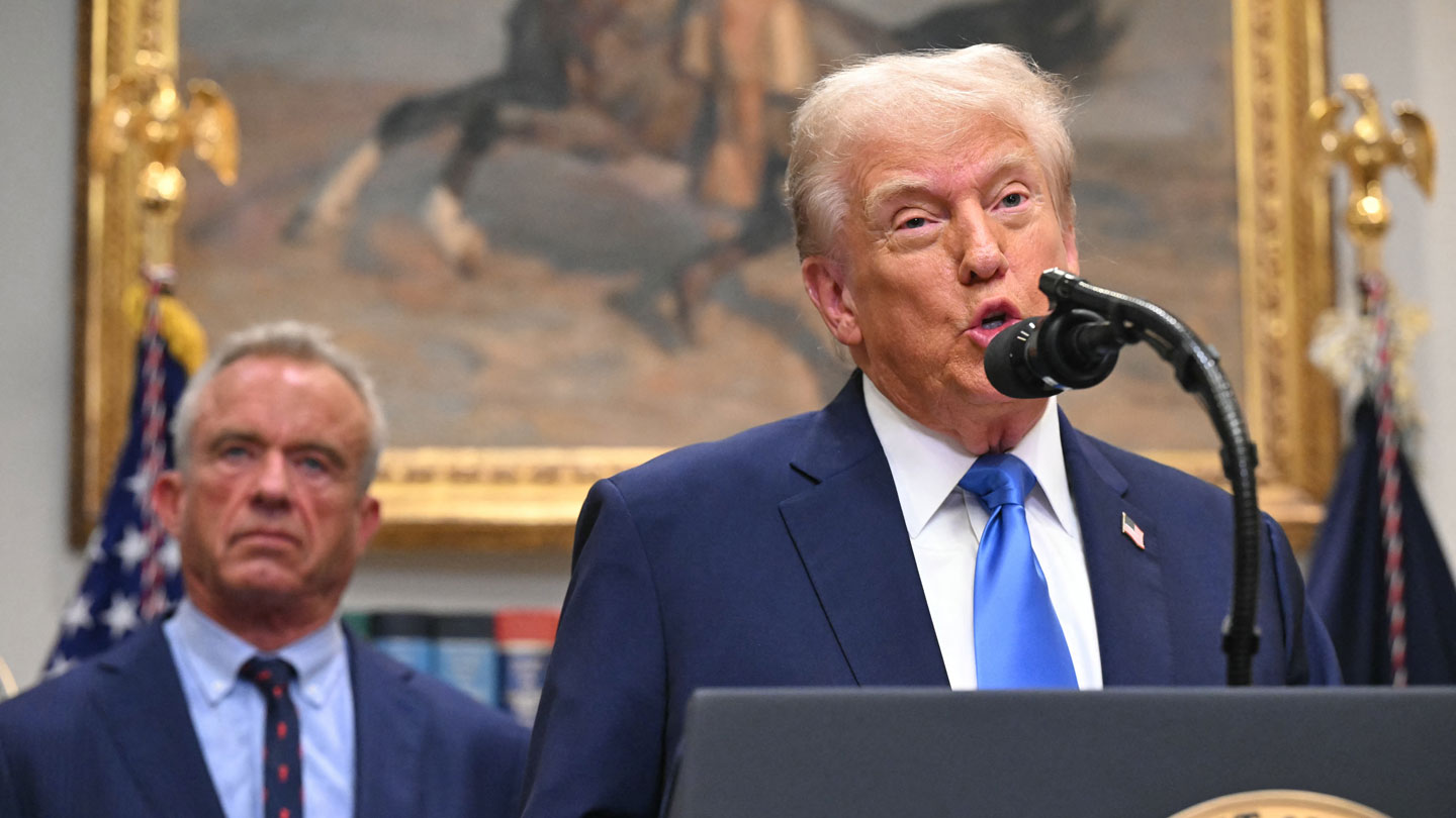 President Trump, in a dark suit and blue tie, speaks into a microphone as Robert F. Kennedy, Jr. stands behind him.