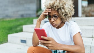 A young Black woman with light-colored hair sitting on stairs looks at her phone. She has a furrowed brow and her lips are pursed as if in worry or confusion. Her right hand is on her brow and her left holds a phone.