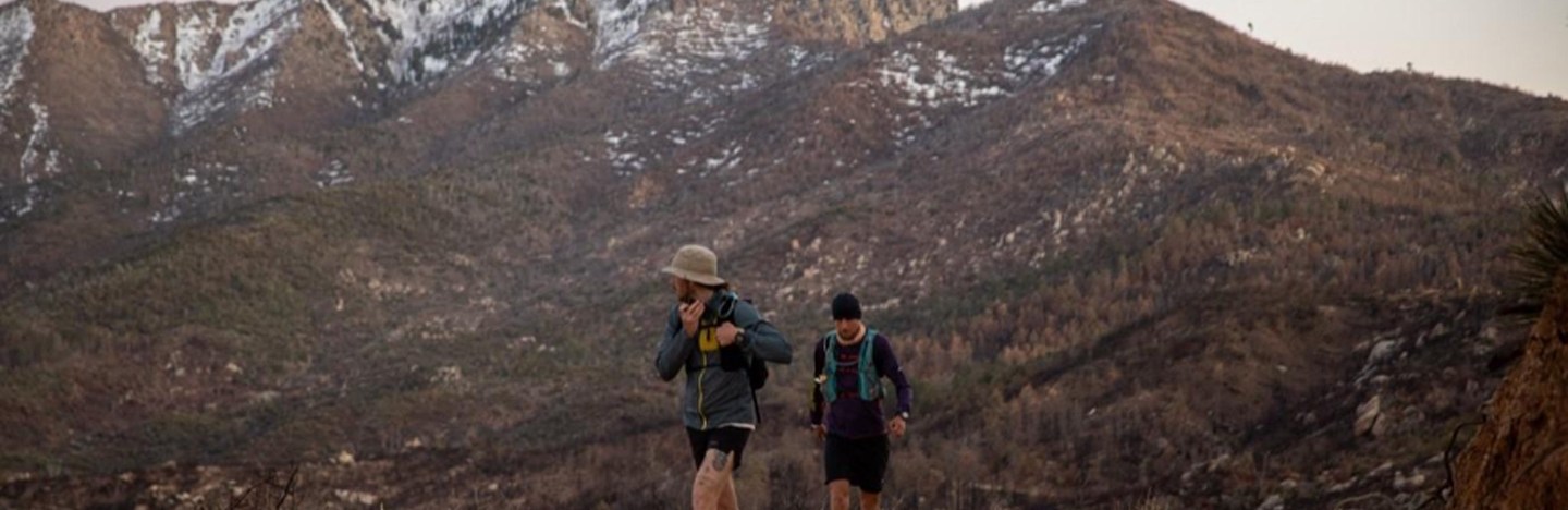 Two men wearing backpacks move along a trail. Mountains with a dusting of snow fill the background.
