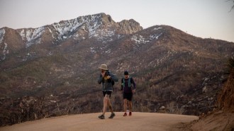 Two men wearing backpacks move along a trail. Mountains with a dusting of snow fill the background.