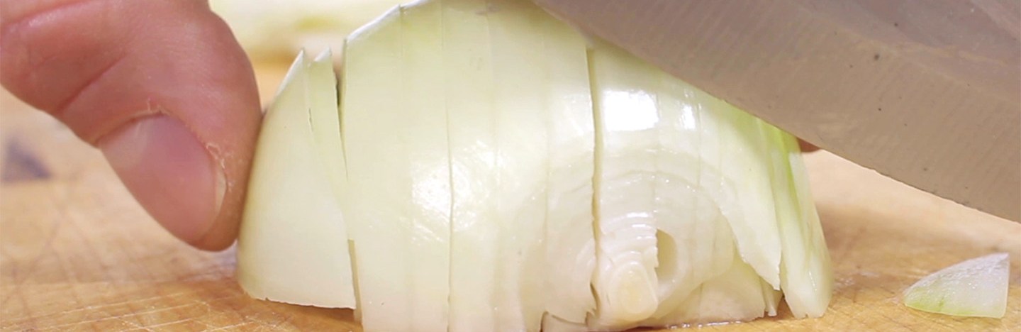 A person holds a knife parallel to a yellow onion cut into horizontal slices. The knife is about to slice the onion in the vertically to dice it into small chunks.