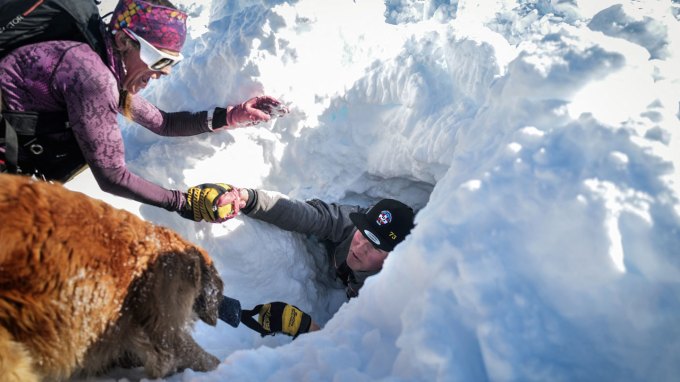 An avalanche rescuer and a rescue dog help pull a person out of a deep pile of snow for a training exercise.