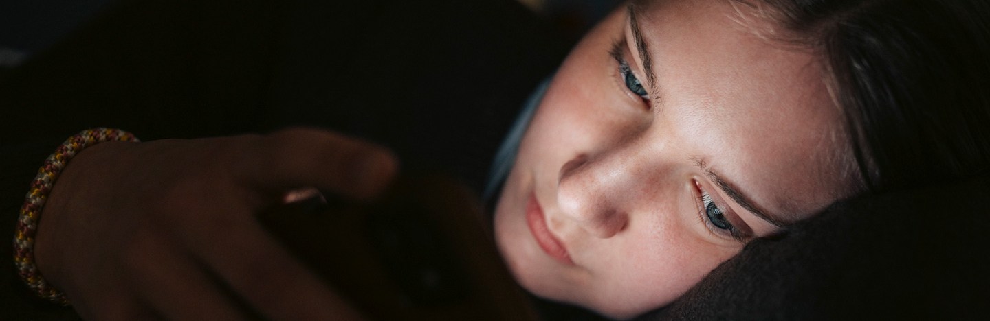 A teenage girl lies on her side in the dark, her face illuminated by her smartphone, suggesting she is interacting with something on the screen.