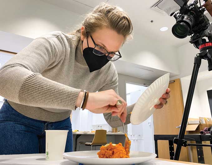 A photograph of a maked researcher pitting food on a plate with a camera trained on the food