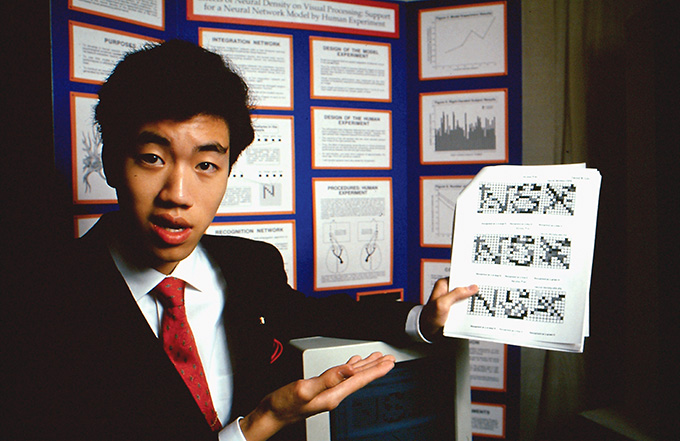 A young David R. Liu stands in front of a tri-fold presentation board in a sport coat and red tie. He holds up a diagram and gestures to it with his right hand. He looks straight into the camera.