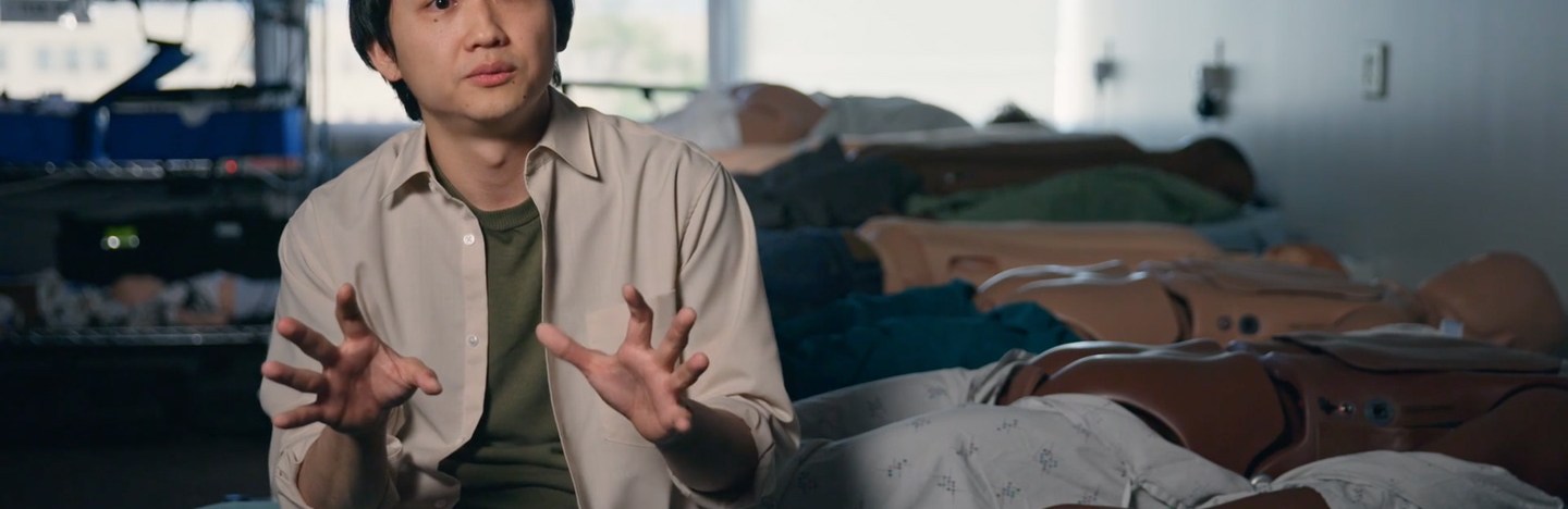 A picture of a man sitting on the edge of a hospital bed in front of a row of mannequins that have their rears exposed