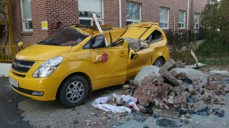 A yellow van crushed by fallen bricks and debris outside a brick building