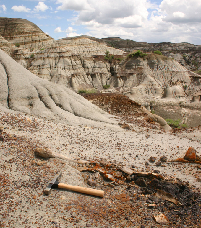 A geological hammer lying on eroded badlands terrain with layered sedimentary rock formations and a cloudy sky in the background.