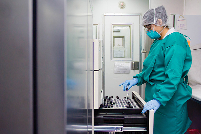 Profile view of a scientist wearing a green lab smock, face mask, hairnet, latex gloves and glasses. The scientist holds a test tube above a an open cabinet drawer inside a lab.