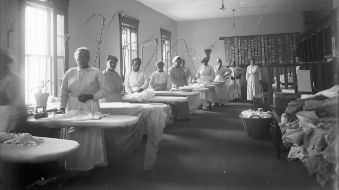 Black women stand at ironing boards in a laundry room at a hospital in 1918.