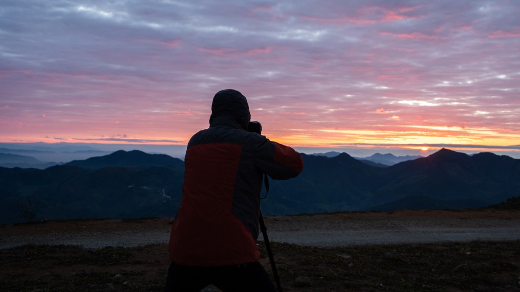 A photographer stands silhouetted against a colorful sunset with low-lit mountains in the distance.
