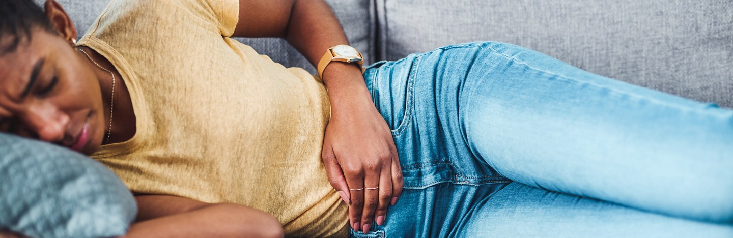 A woman lies on her side on a couch, holding her stomach. She has a look of discomfort on her face.