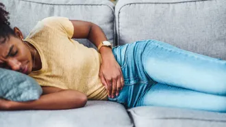 A woman lies on her side on a couch, holding her stomach. She has a look of discomfort on her face.