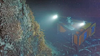 Equipment with bright lights sits on the seafloor in dark murky water, its lights trained on a hydrothermal vent rising up beside it.