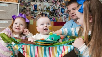 A smily chubby-cheeked baby is sitting in a rainbow striped cloth baby seat. His two sisters and brother sit around him. Everyone is smiling.