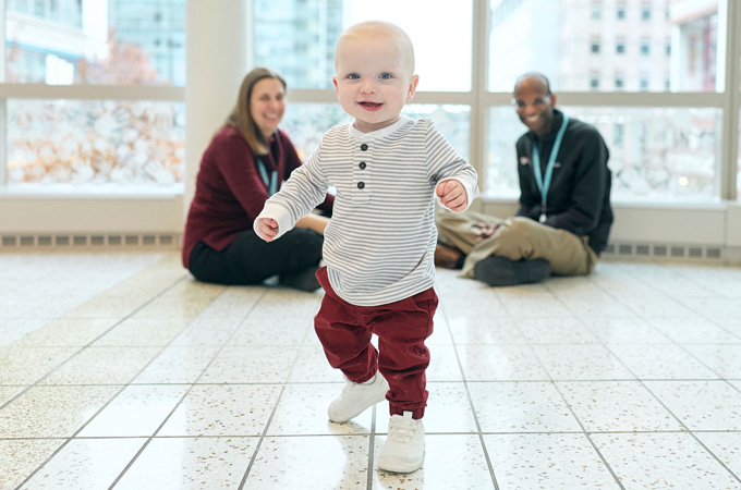 A little boy walks toward the camera. He's wearing red pants, a beige and white striped top and big smile. Two adults sit in the background smiling.
