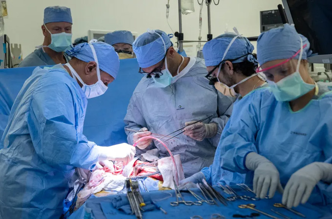 Six medical professionals wearing masks, gloves and protective coverings surround an opening in sheets covering a patient. They are performing surgery.