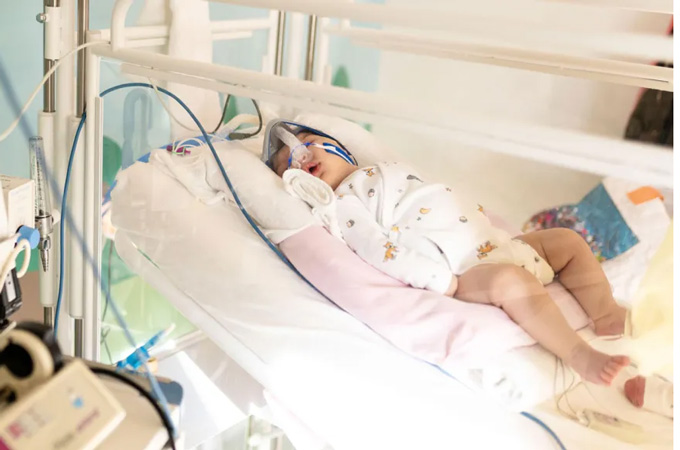A baby lies in a hospital bed attached to oxygen and other tubes.