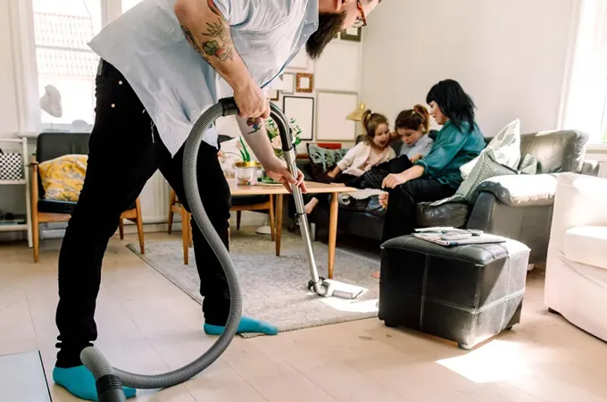 A man vacuums while a woman sits on a couch reading with two children.