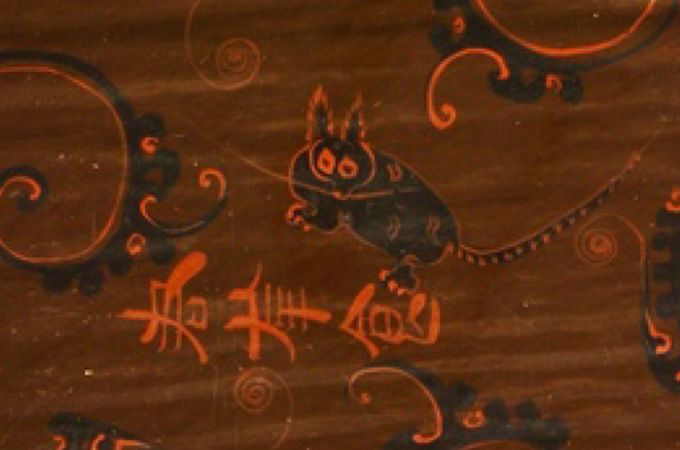 A design on an artifact includes a drawing of what looks like a black cat with pointy fuzzy ears, pointy claws, spots on its back and stripes on its tail.