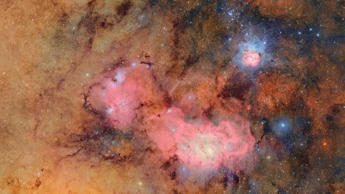 This cosmic image of two nebulas shows a bright pink splotch in the middle and lots of points of light speckling the whole scene.