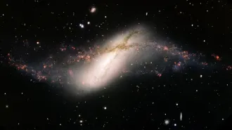 A telescope image shows a bright, elongated galaxy with a glowing central core encircled by a warped ring of stars and gas set against a field of stars and galaxies.