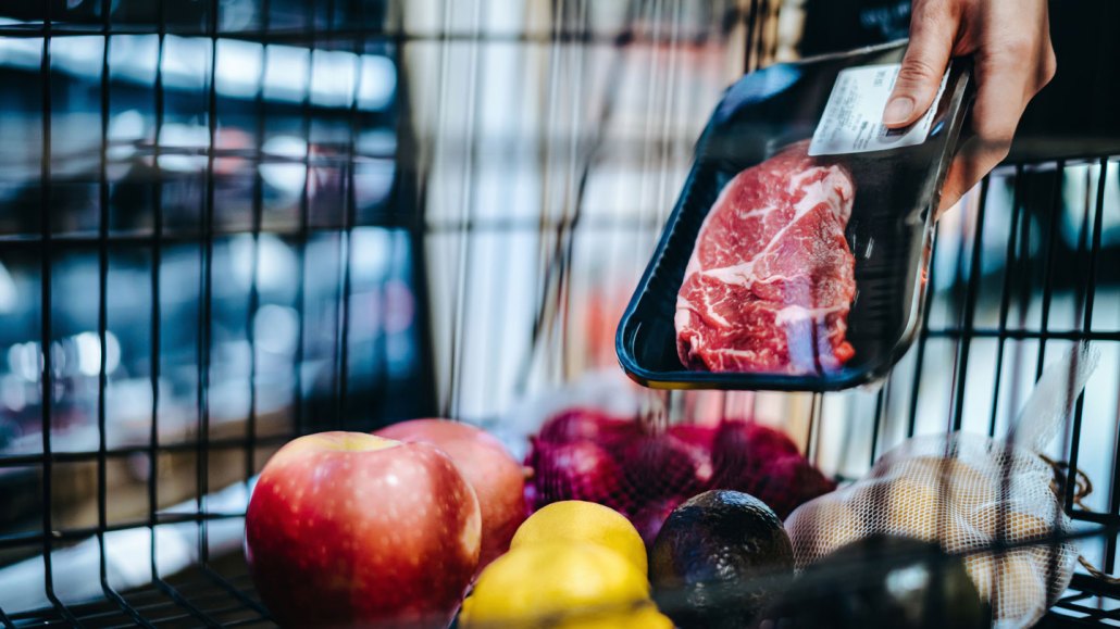 A person placing a piece of meat, plastic wrapped in a black tray, into a basket full of fruits and vegetables.