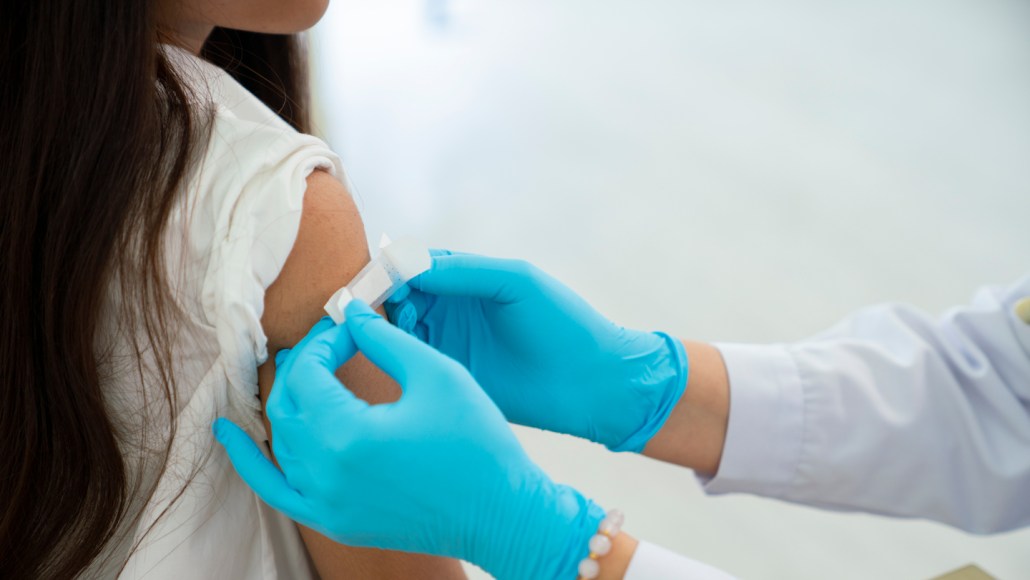 A health care worker wearing blue gloves applies a small adhesive bandage to a child’s upper arm after vaccination.