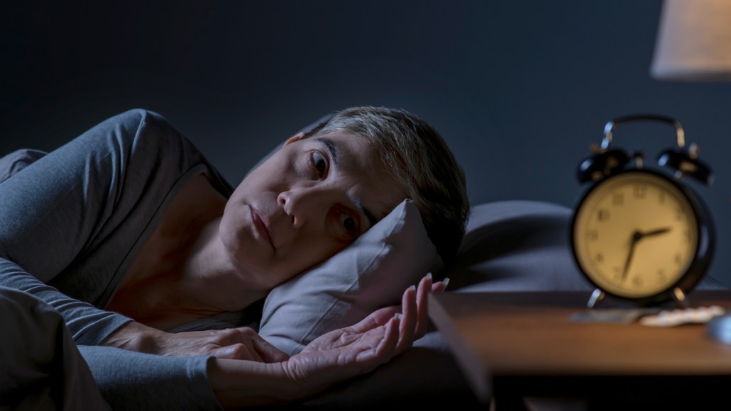 An older woman lays on her side in bed and stares at her alarm clock