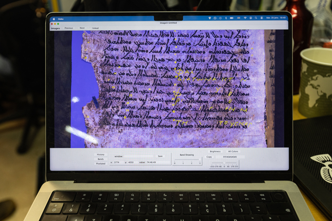 A laptop screen displays a digitally enhanced image of an ancient manuscript with handwritten text highlighted.