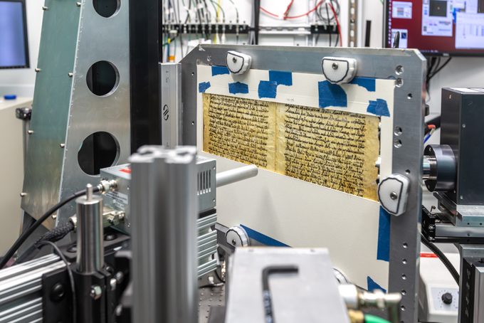 A fragile manuscript page is mounted in a metal frame inside a laboratory imaging system surrounded by scientific equipment.