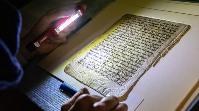 A researcher shines a handheld light across fragile parchment containing handwritten text.