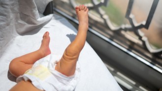 A diapered baby is shown from the torso down laying in front of a window with sunlight streaming in