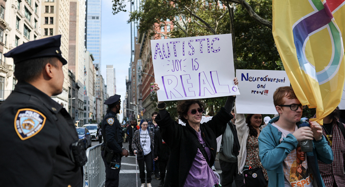 A person holds a sign that says "Autistic joy is real" as they walk past a police officer in New York City.