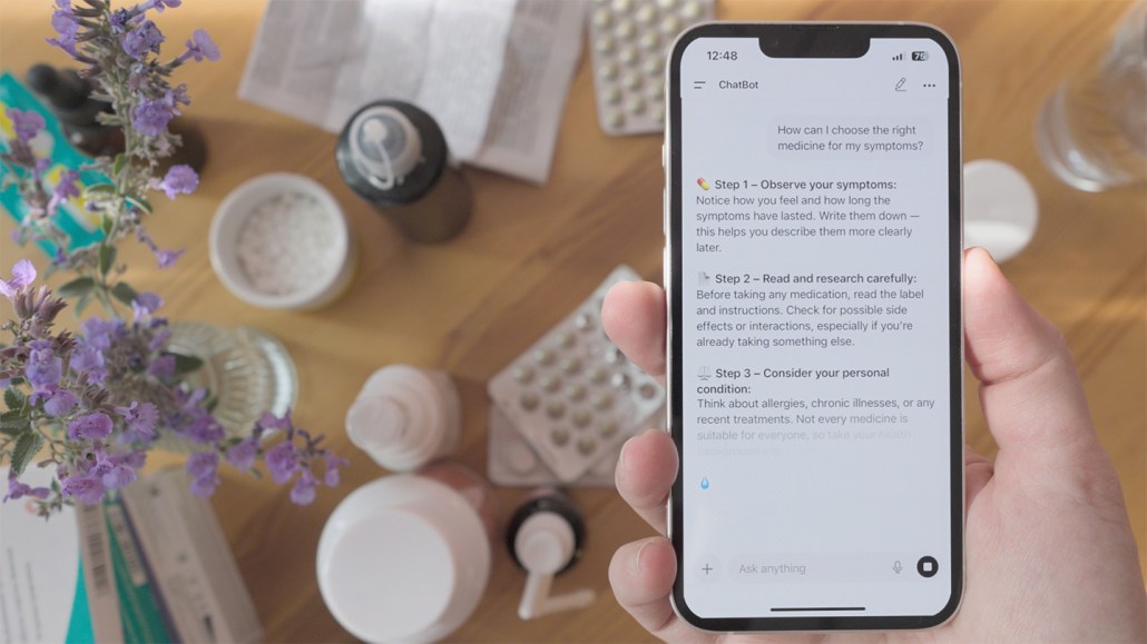 A hand holds a phone. The screen shows part of a conversation with a chatbot about medical questions. Pills, bottles, flowers and a glass of water are in the background.