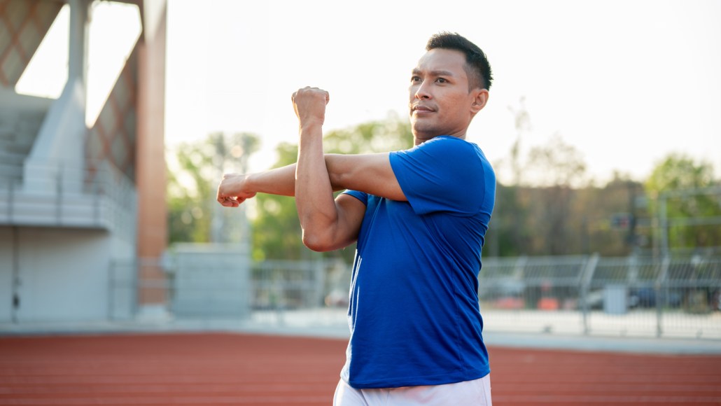 A man in a blue shirt stands on an outdoor track stretching his shoulder.