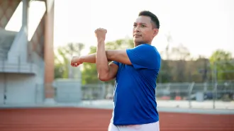 A man in a blue shirt stands on an outdoor track stretching his shoulder.