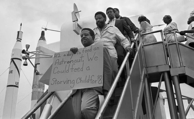 Four people stand on stairs. One of them holds a sign. A space rocket can be seen in the background.