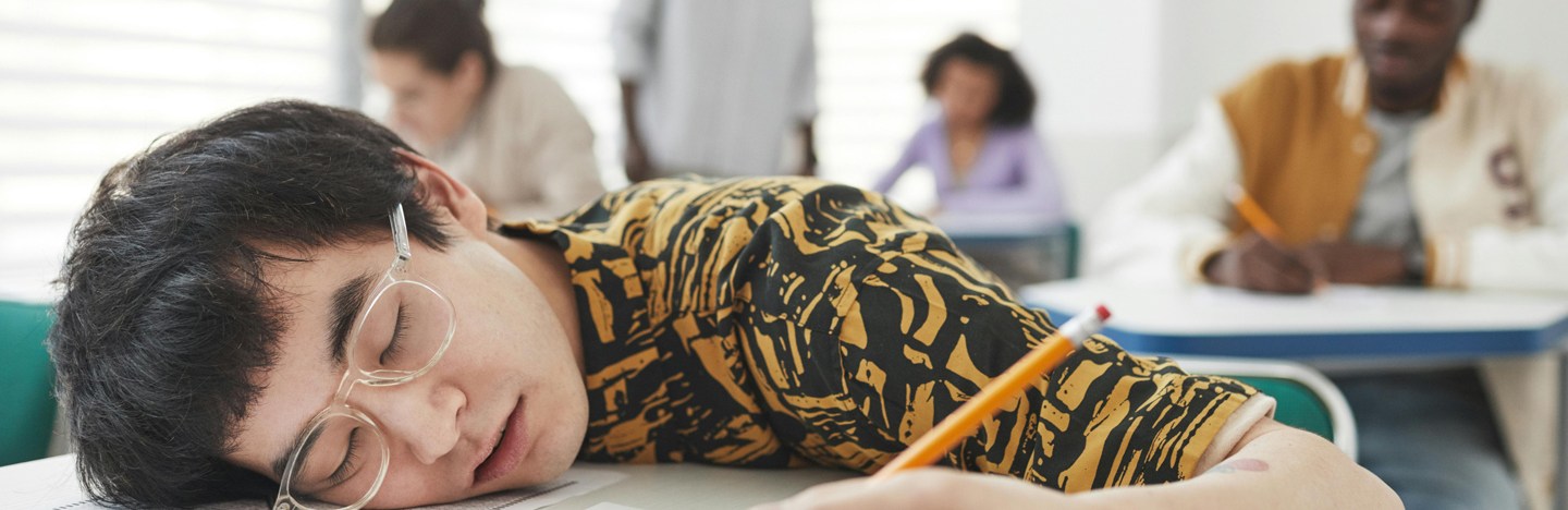 A high school student is sleeping on their desk, which has papers. They are holding a pencil.