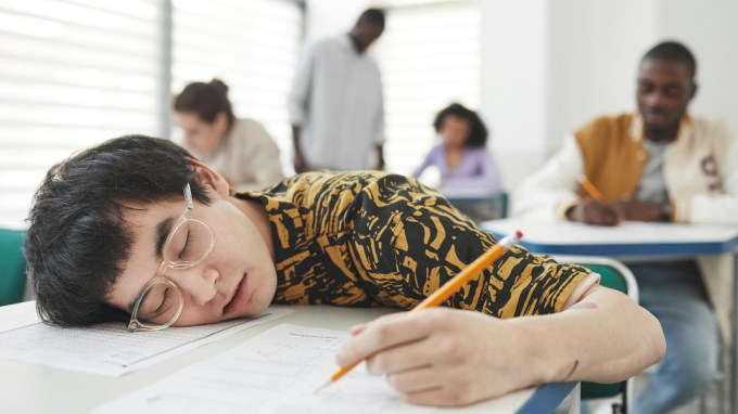 A high school student is sleeping on their desk, which has papers. They are holding a pencil.