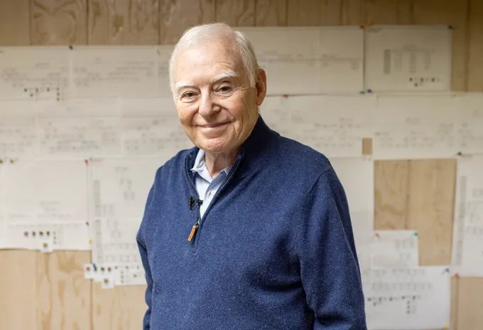 A white-haired man wearing a blue sweater smiles.