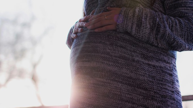 A pregnant woman wearing a sweater places her hands on her belly.