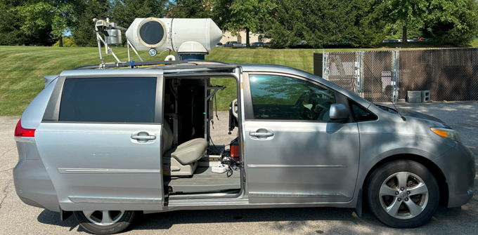 A silver 2013 Toyota Sienna van with the sliding back doors open. The van has been modified with a cylindrical piece of equipment mounted on the roof. The van is parked in a concrete parking lot in front of grass trees, and a fenced off area.